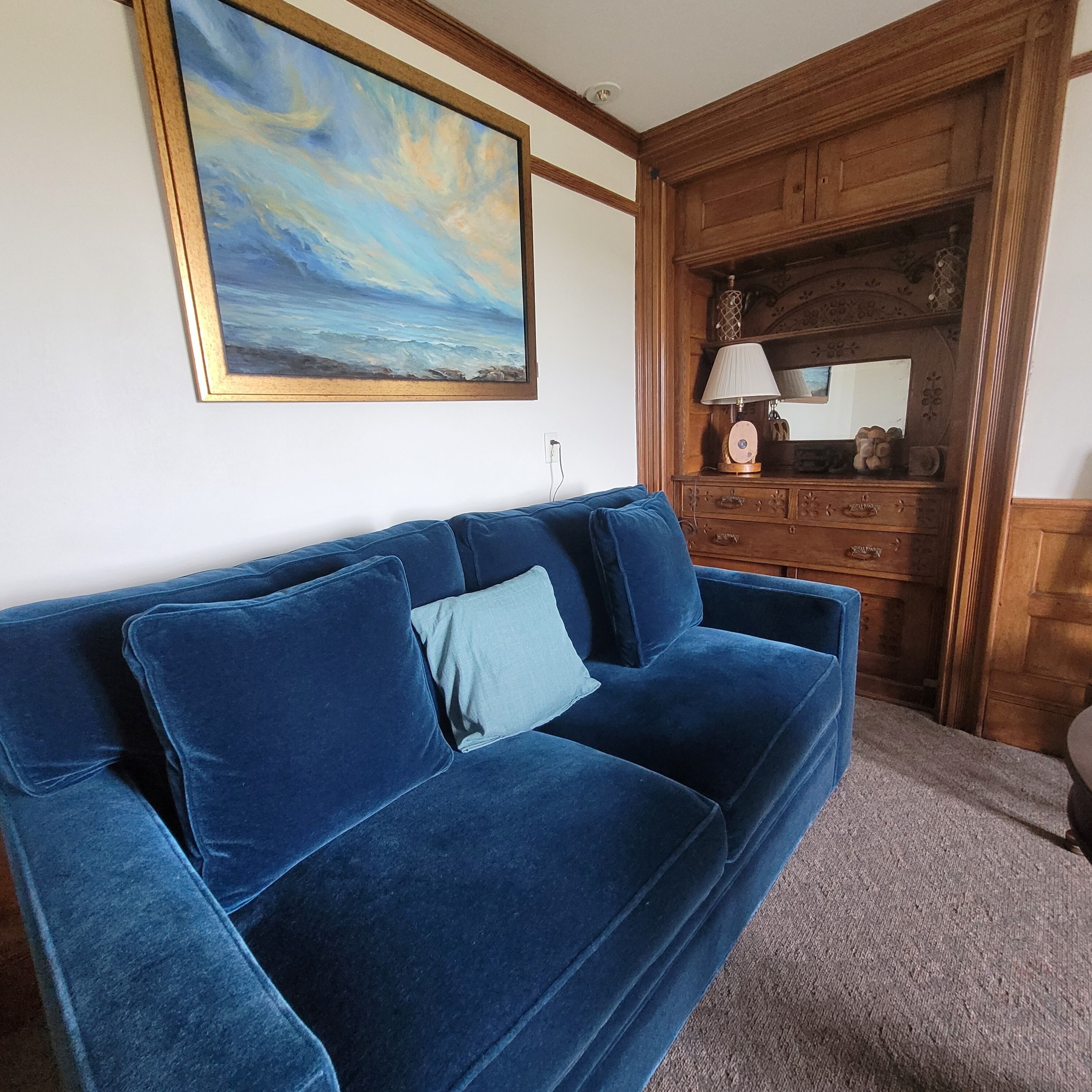 Un canapé en velours bleu avec un coussin bleu clair, un meuble en bois avec un miroir, et un tableau représentant un ciel bleu accroché au mur.