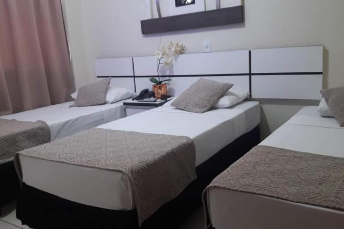 A hotel room with three single beds, each with a patterned bedspread and pillows. A small table with a phone and a vase of flowers is between the beds.