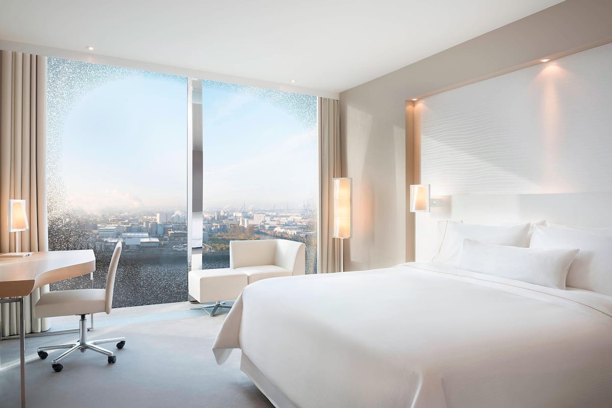 Une chambre d’hôtel moderne avec un grand lit, un bureau et une chaise. Une grande fenêtre offre une vue sur la ville.