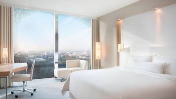 Une chambre d’hôtel moderne avec un grand lit, un bureau et une chaise. Une grande fenêtre offre une vue sur la ville.