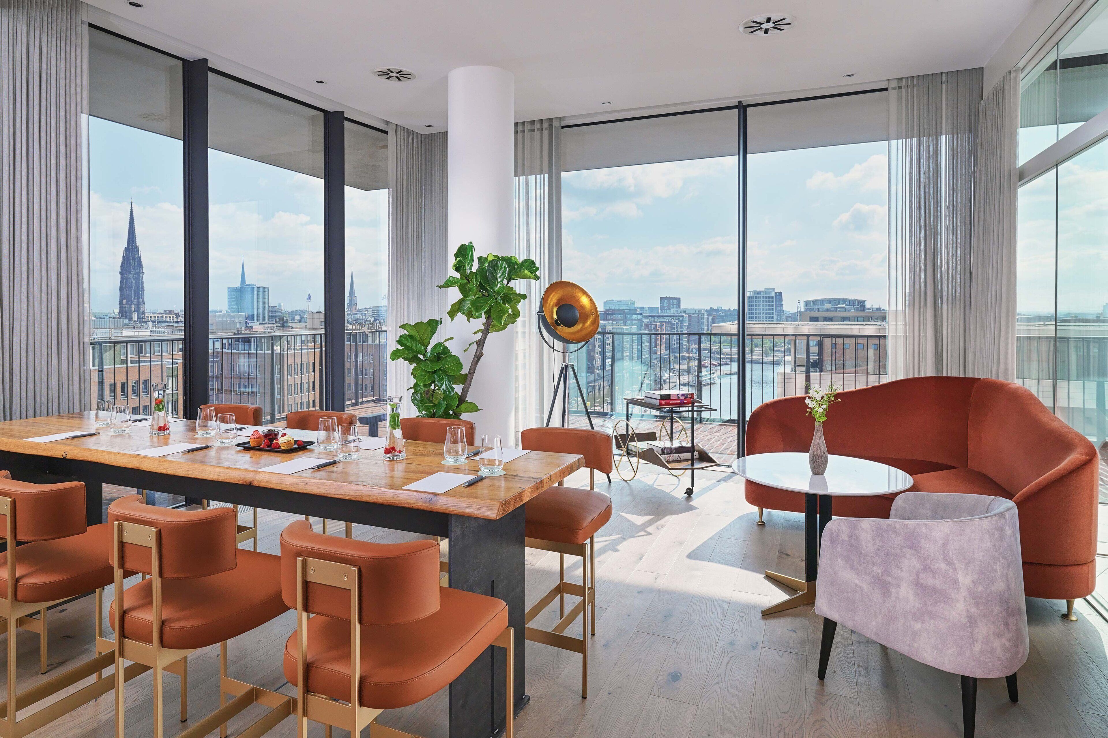 Una habitación de hotel moderna con una mesa de comedor grande, sillas anaranjadas, una mesa redonda con una silla morada y vistas al horizonte de la ciudad.