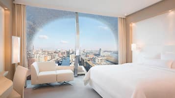 Camera d'albergo moderna con un'ampia finestra che offre vista sulla città, un letto, un angolo salotto e una scrivania.