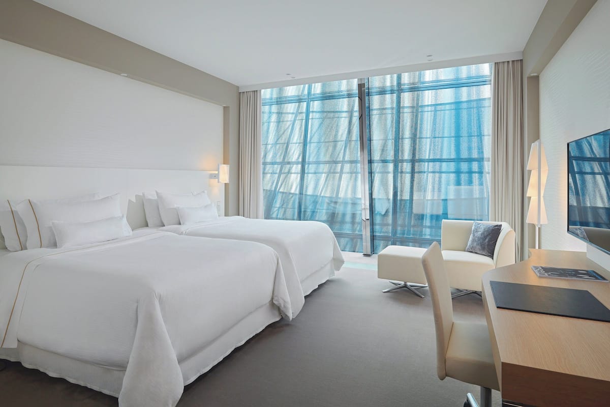 Une chambre d’hôtel avec deux lits, un bureau, une chaise et une grande fenêtre avec des rideaux.