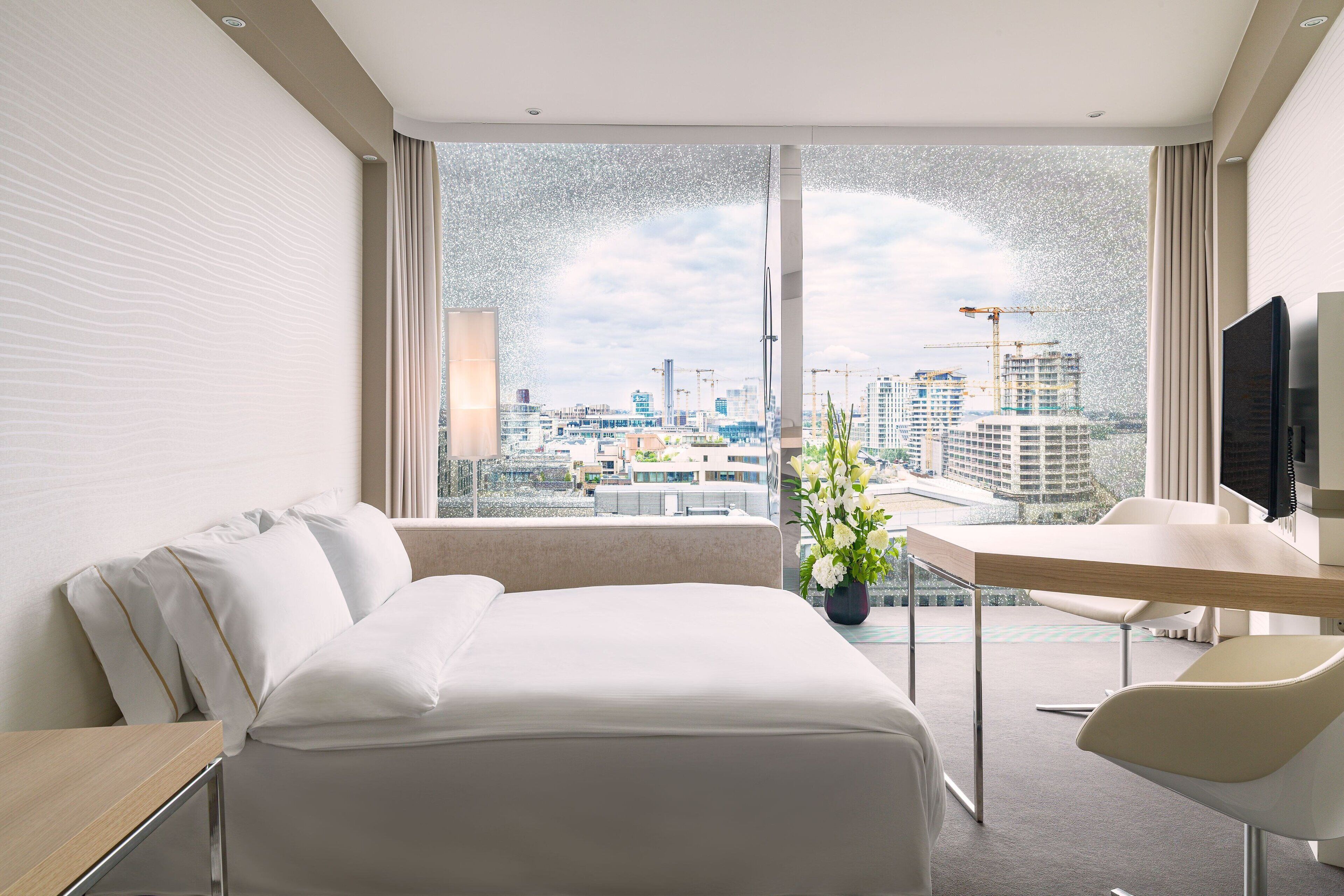 Ein Hotelzimmer mit Bett, Schreibtisch, Sessel, Fernseher und großem Fenster mit Blick auf die Stadt.