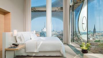 A modern hotel room with a large bed, a bedside table with a lamp, a remote control, and a view of a cityscape through a large window.