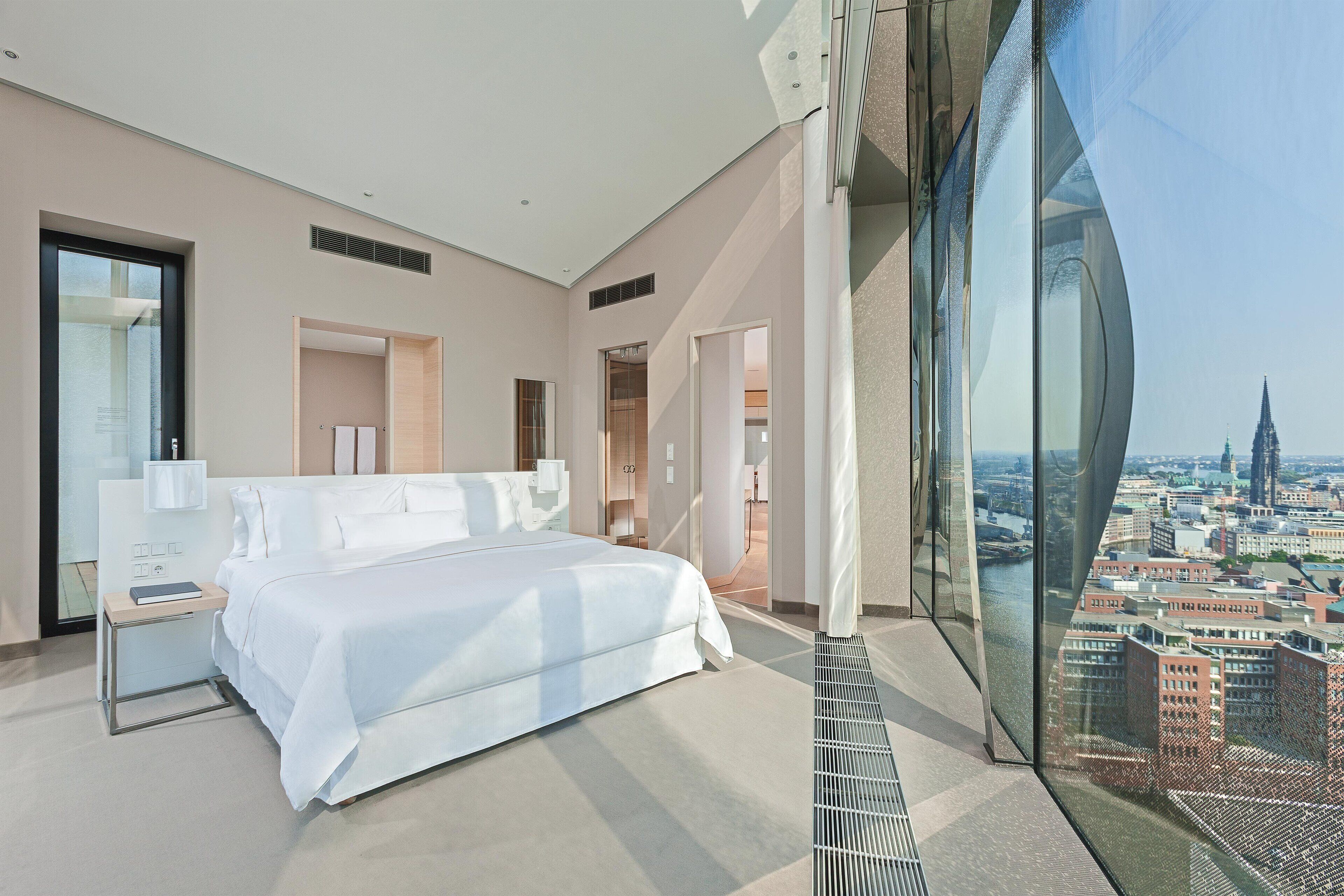 Ein modernes Schlafzimmer mit einem großen Bett, einem Nachttisch und Blick auf die Stadt durch raumhohe Fenster.