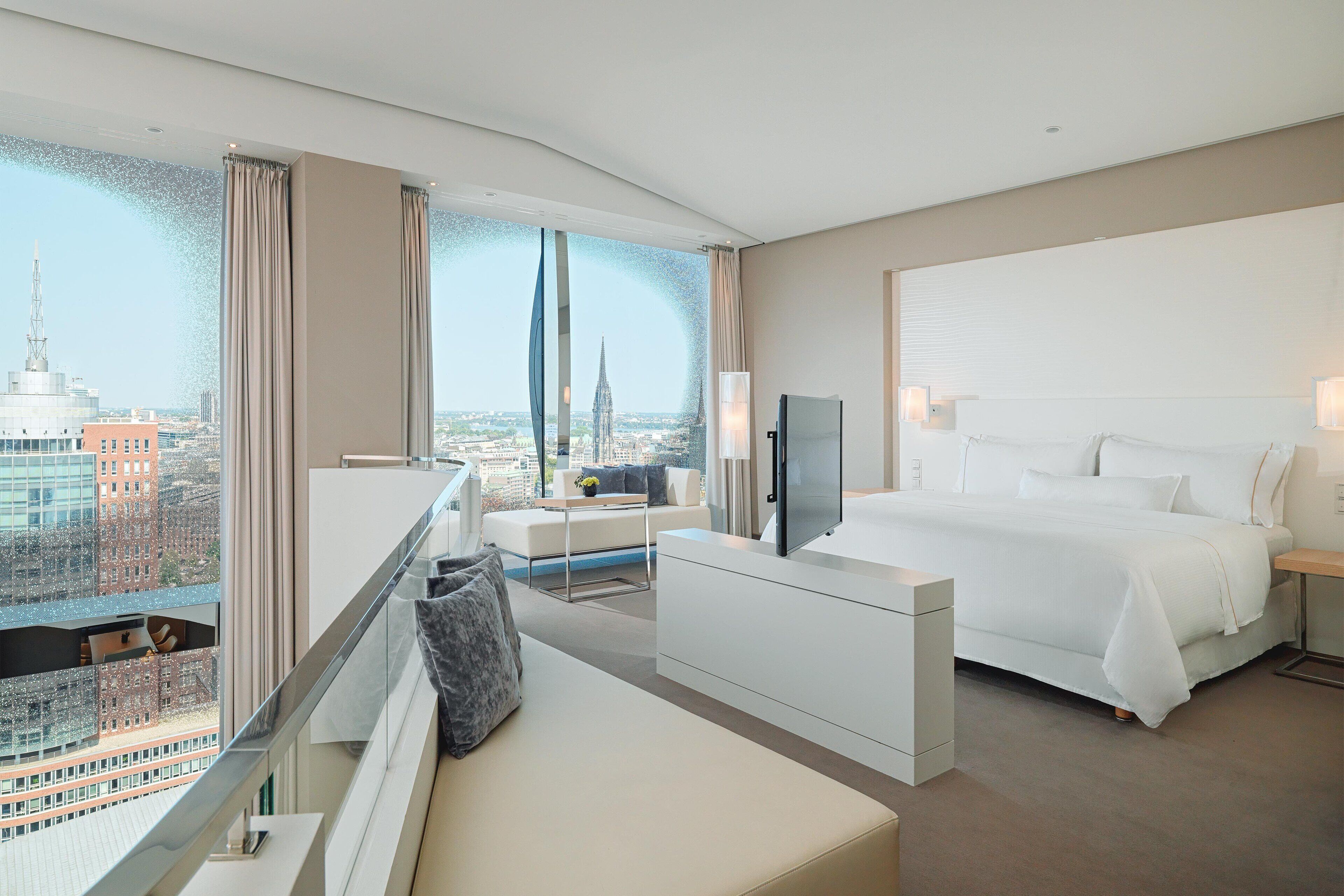 A modern hotel room with a large bed, a desk, and a seating area. The room has a city view through large windows.