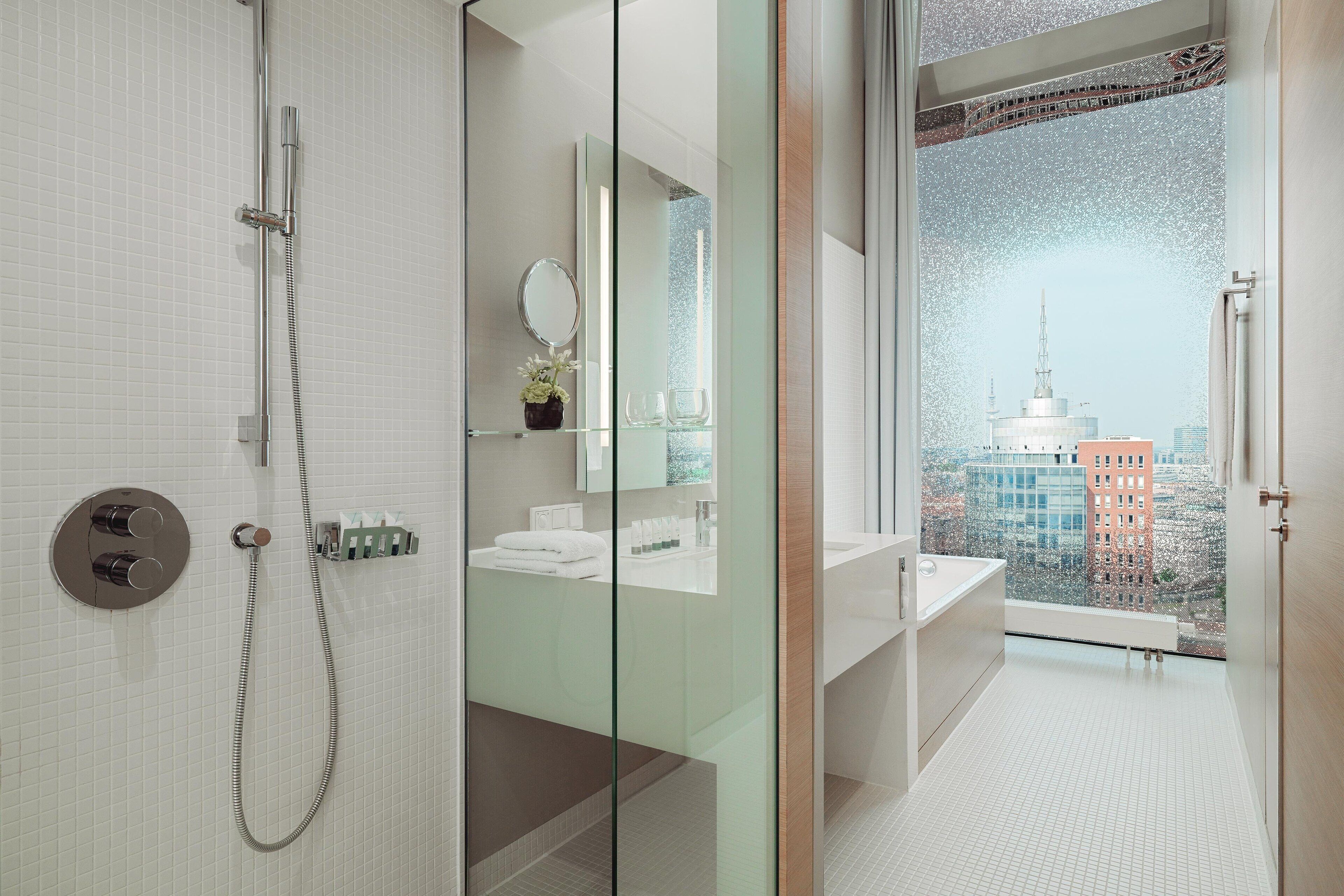 A modern bathroom with a glass shower enclosure, a large mirror, and a view of a cityscape through a large window.