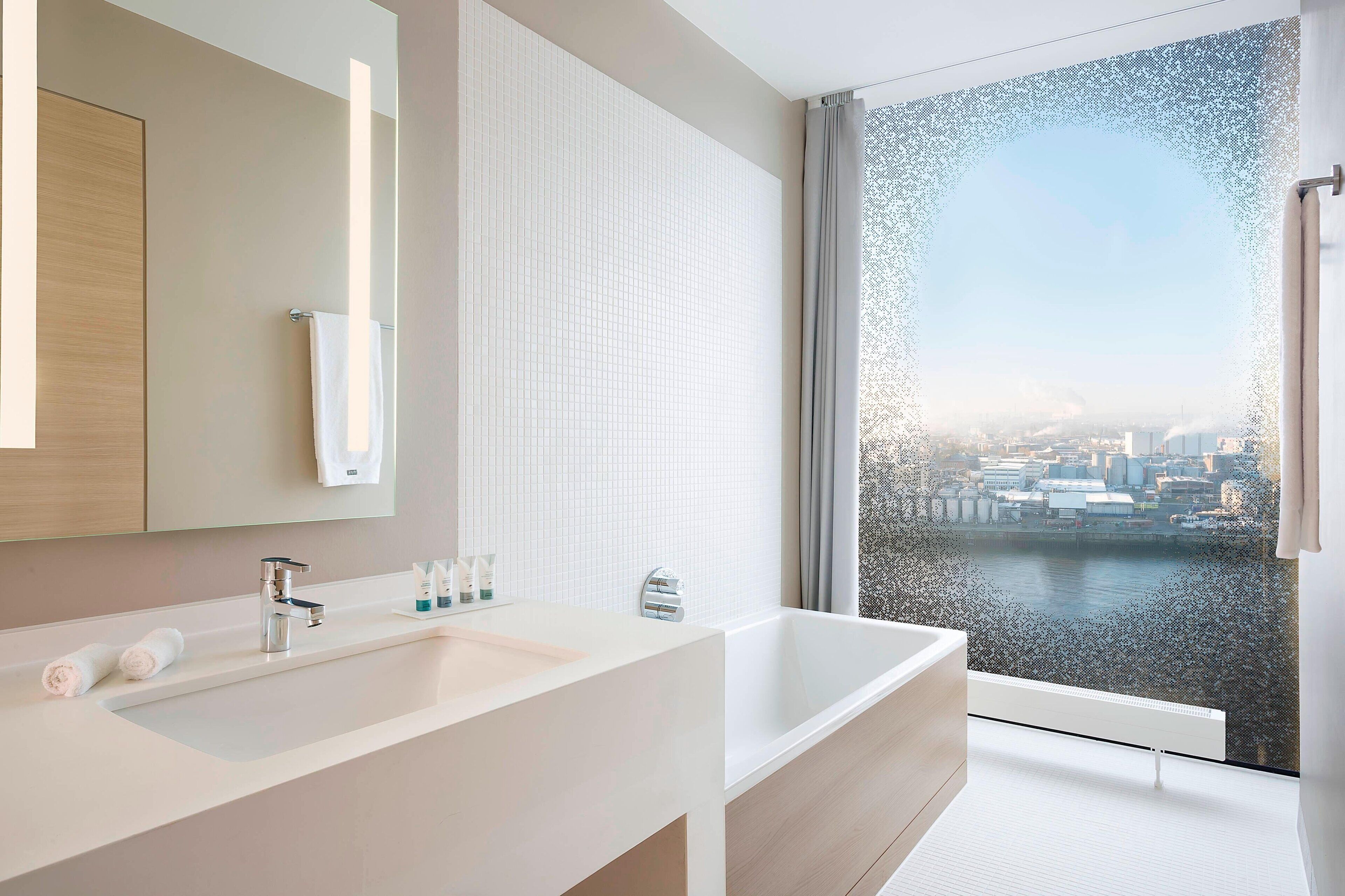 A modern bathroom with a large bathtub, a white sink, and a mirror. There is a view of a cityscape through a large window.