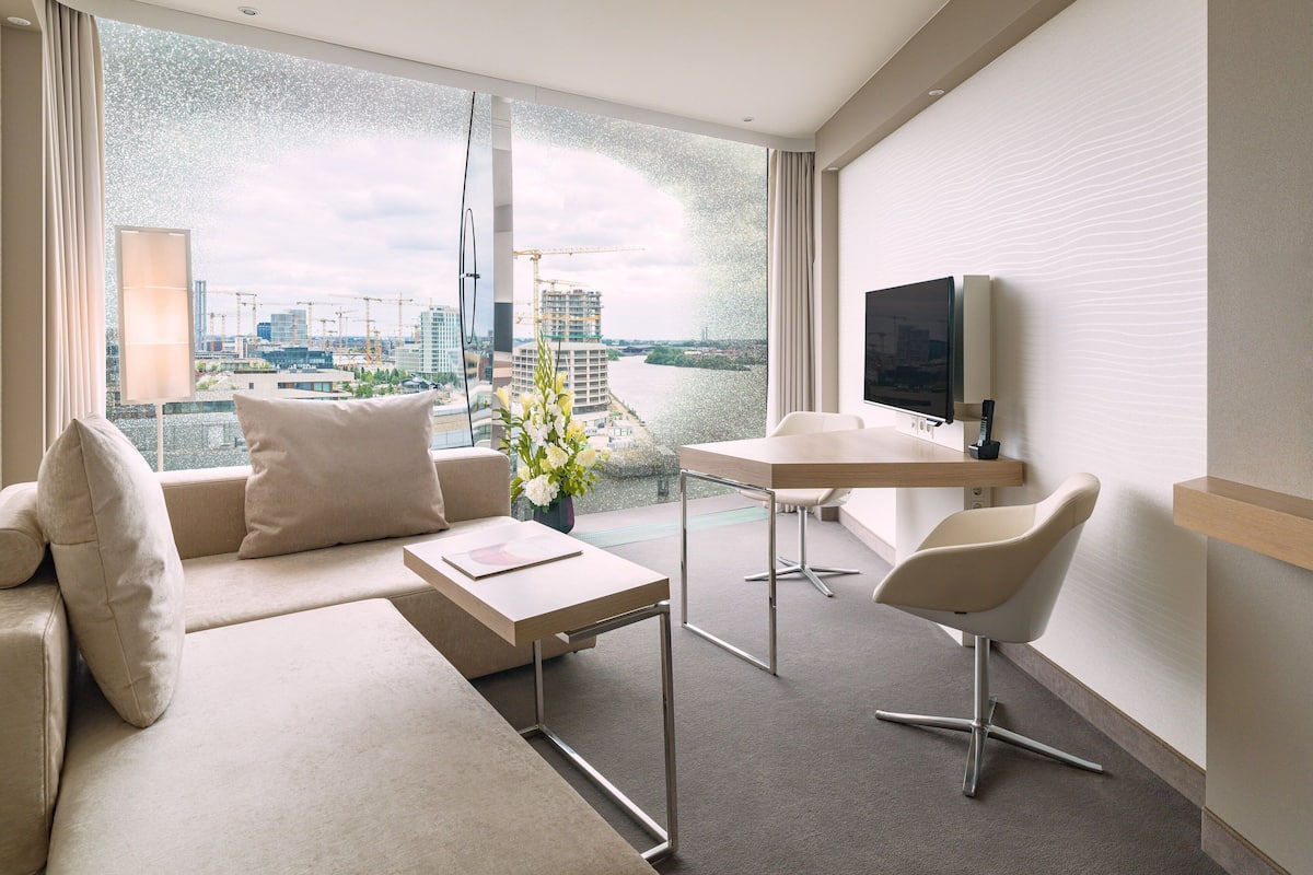 Une chambre d’hôtel moderne dotée d’une grande fenêtre donnant sur un paysage urbain et une rivière. On y trouve un canapé, une petite table et une chaise.
