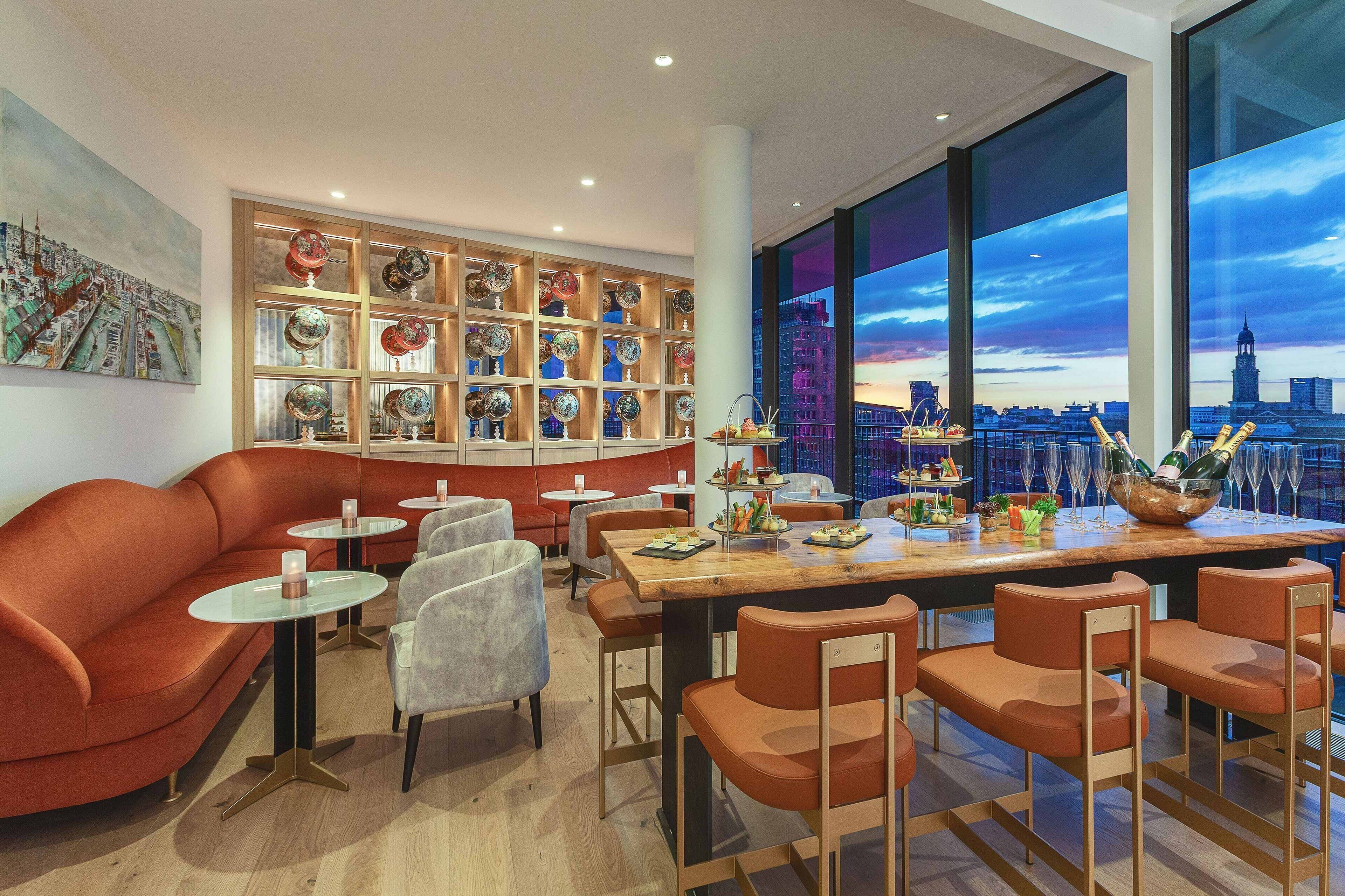 A modern restaurant with a city view, featuring a large dining table, red seating, and a decorative wall with plates.