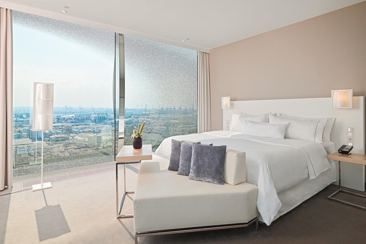 Une chambre d’hôtel moderne dotée d’un grand lit, d’une table de chevet, d’une petite table et offrant une vue sur le paysage urbain grâce à une grande fenêtre.