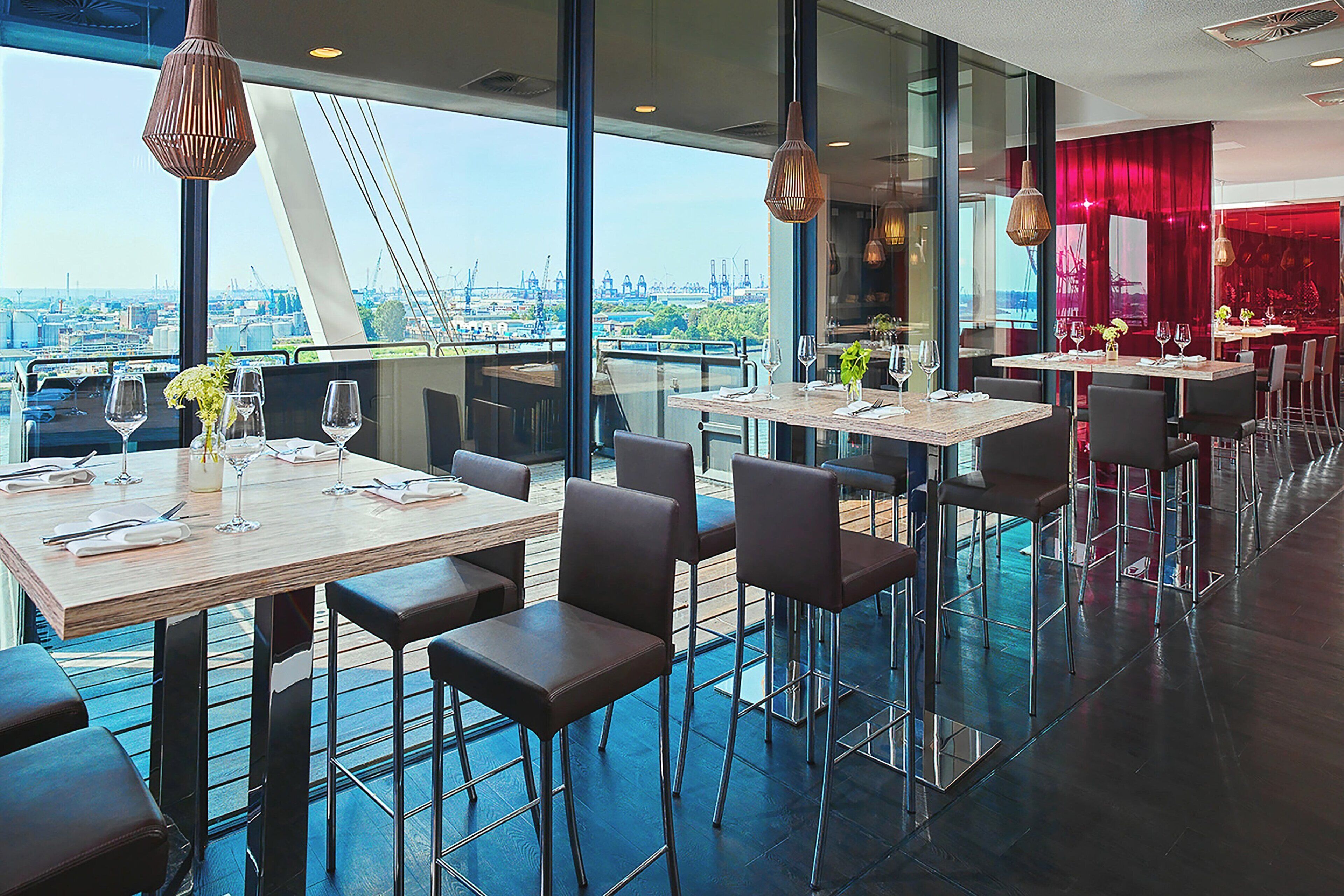 Un restaurante moderno con mesas altas y sillas, ventanas amplias que ofrecen vista a la ciudad y detalles en rojo.