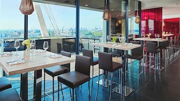 Un restaurante moderno con mesas altas y sillas, ventanas amplias que ofrecen vista a la ciudad y detalles en rojo.