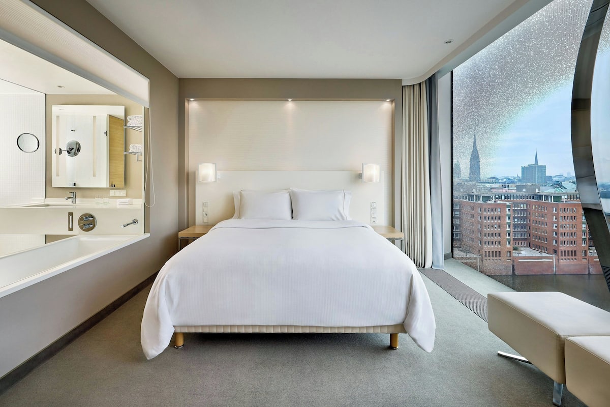 Une chambre d’hôtel moderne avec un grand lit, une salle de bain dotée d’un miroir et d’un lavabo, et une vue sur le paysage urbain.