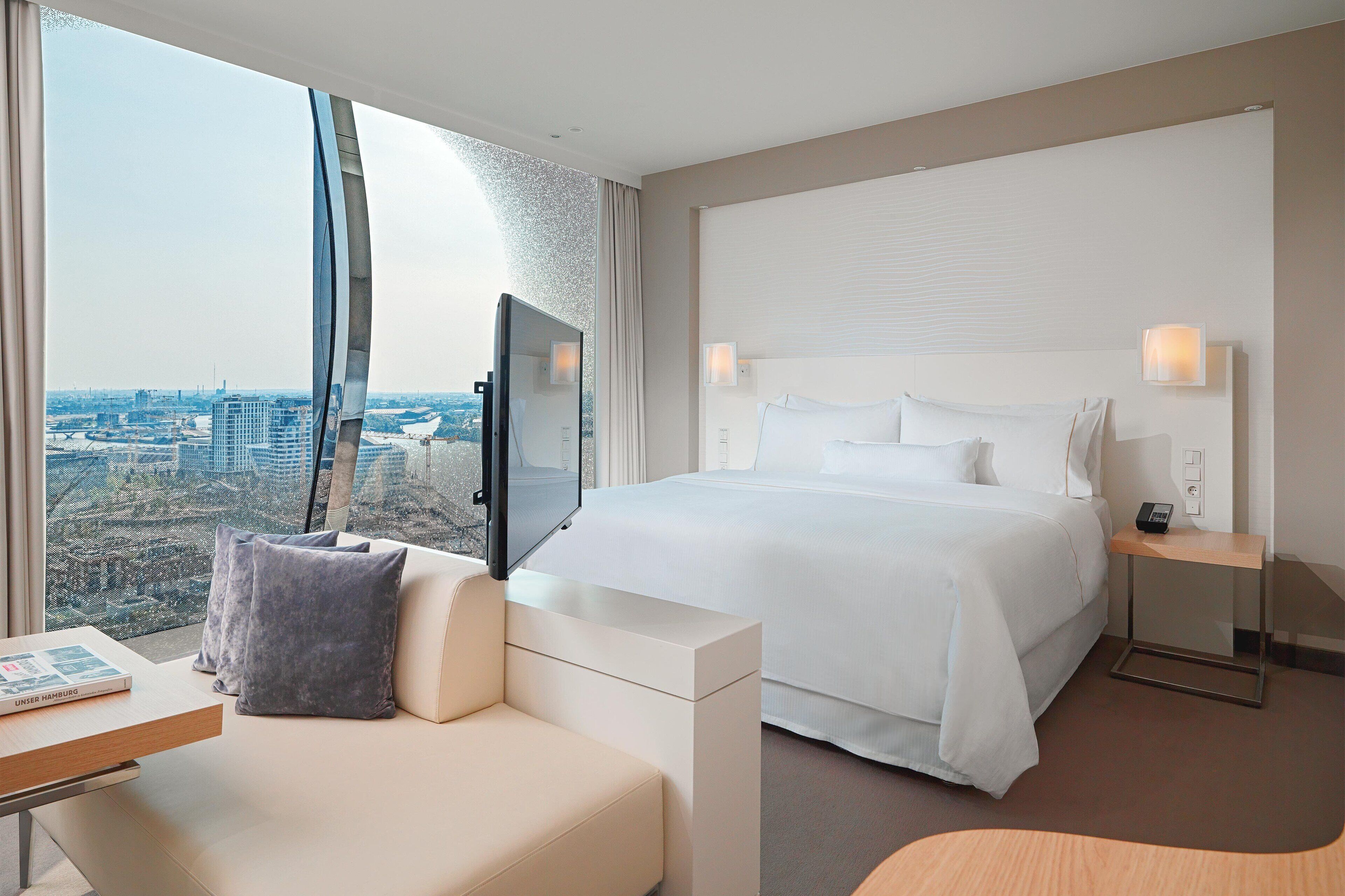 A hotel room with a large bed, a TV, a desk, and a view of the city.