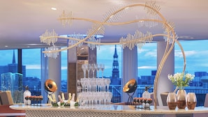 Eine Bar mit einer dekorativen goldfarbenen Struktur, einem Weinkühler und Blick auf eine Stadtlandschaft.