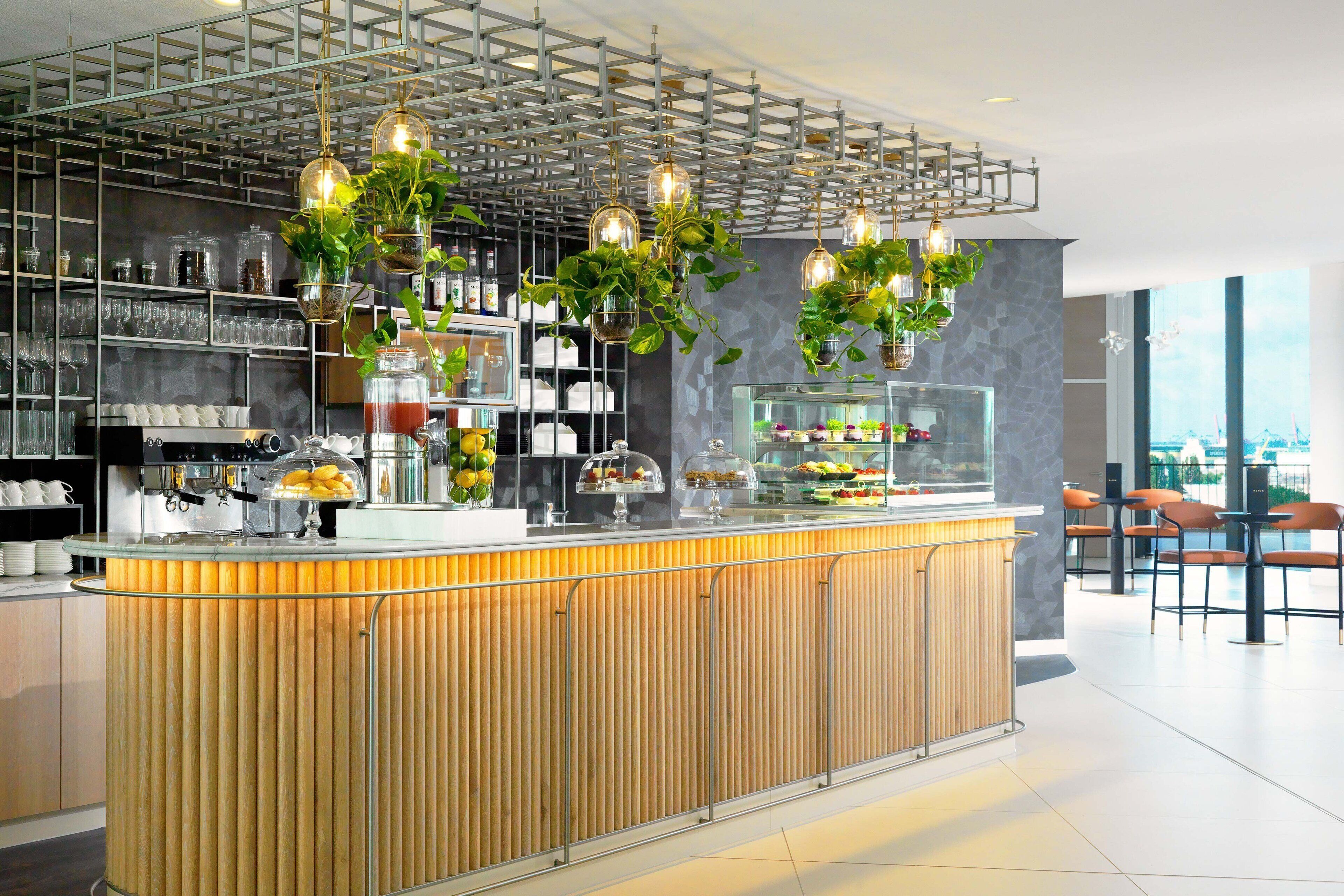 Eine moderne Bar mit Holztresen, hängenden Pflanzen und einer Glastheke mit Lebensmitteln.