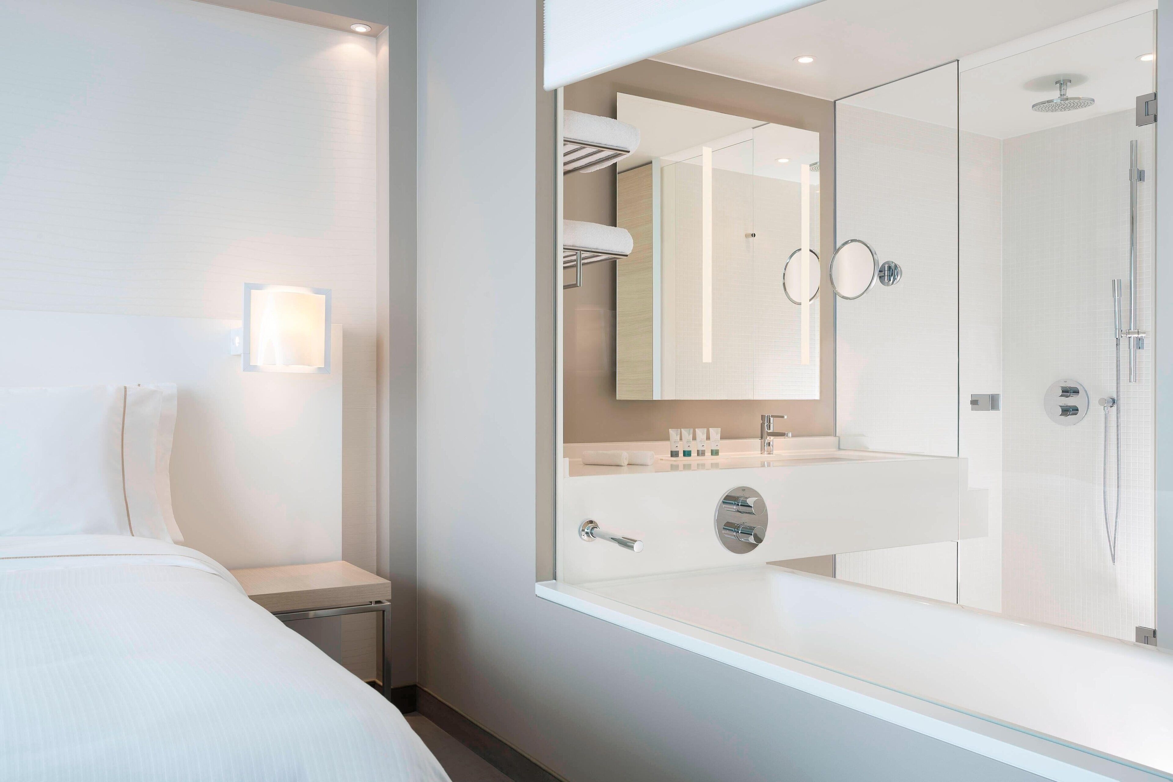 Ein modernes Hotelzimmer mit einem großen Bett, einer Duschkabine aus Glas, einem Schminktisch mit Spiegel und einem Nachttisch.