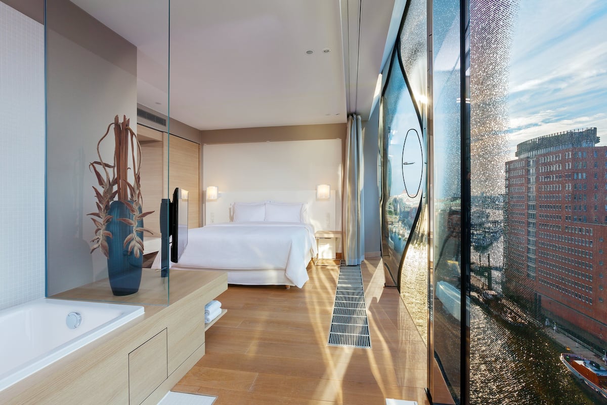Une chambre d’hôtel moderne dotée d’un grand lit, d’une salle de bain avec baignoire et offrant une vue sur le paysage urbain grâce à une grande fenêtre.