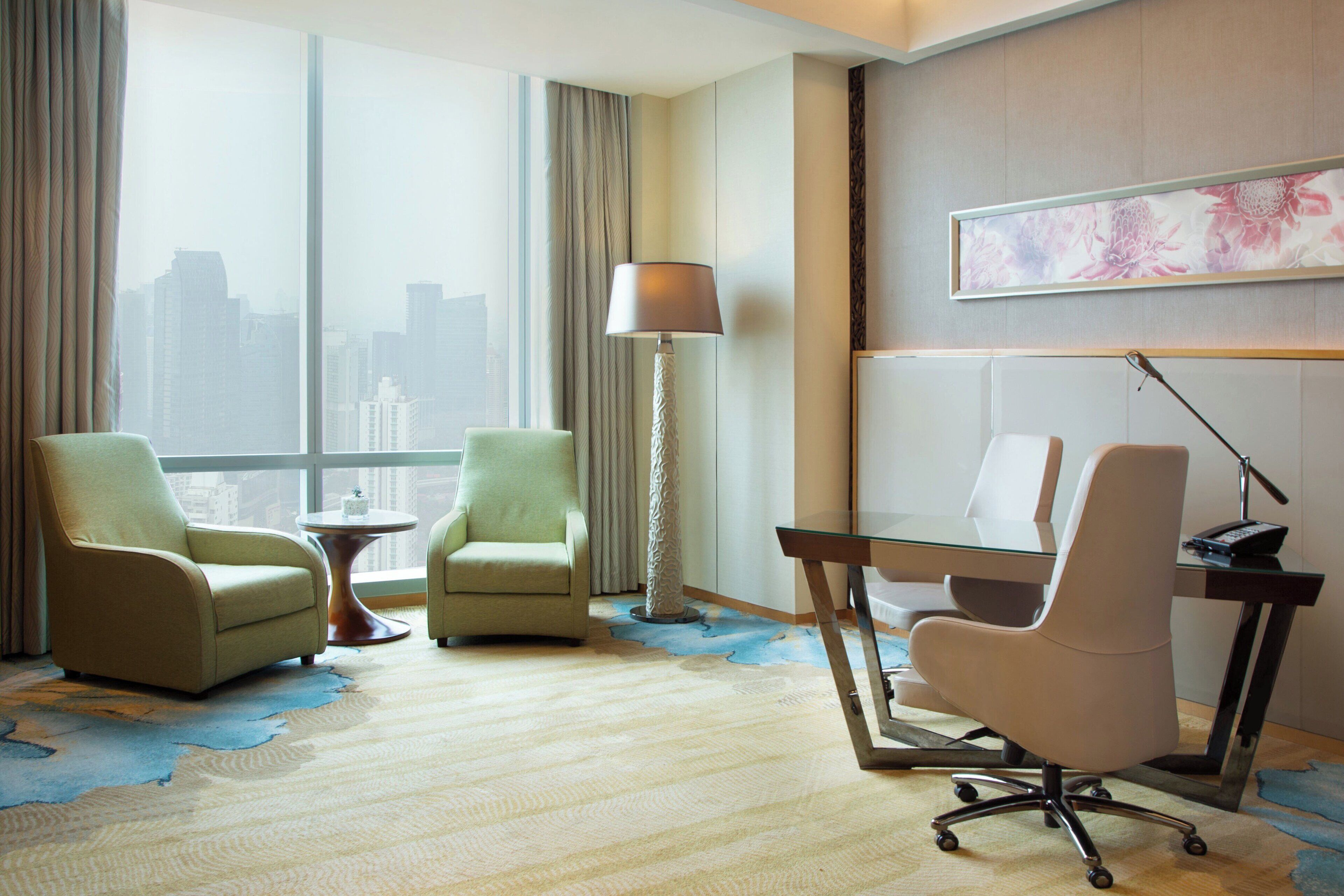 A hotel room with two armchairs, a desk, a lamp, and a large window offering a city view.