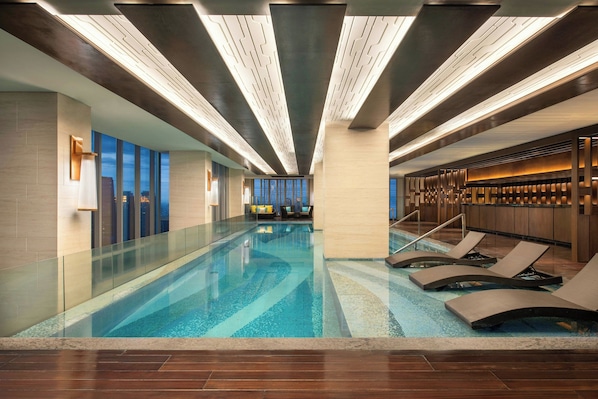 A modern indoor swimming pool with clear water, lounge chairs, and a view of the city through large windows.