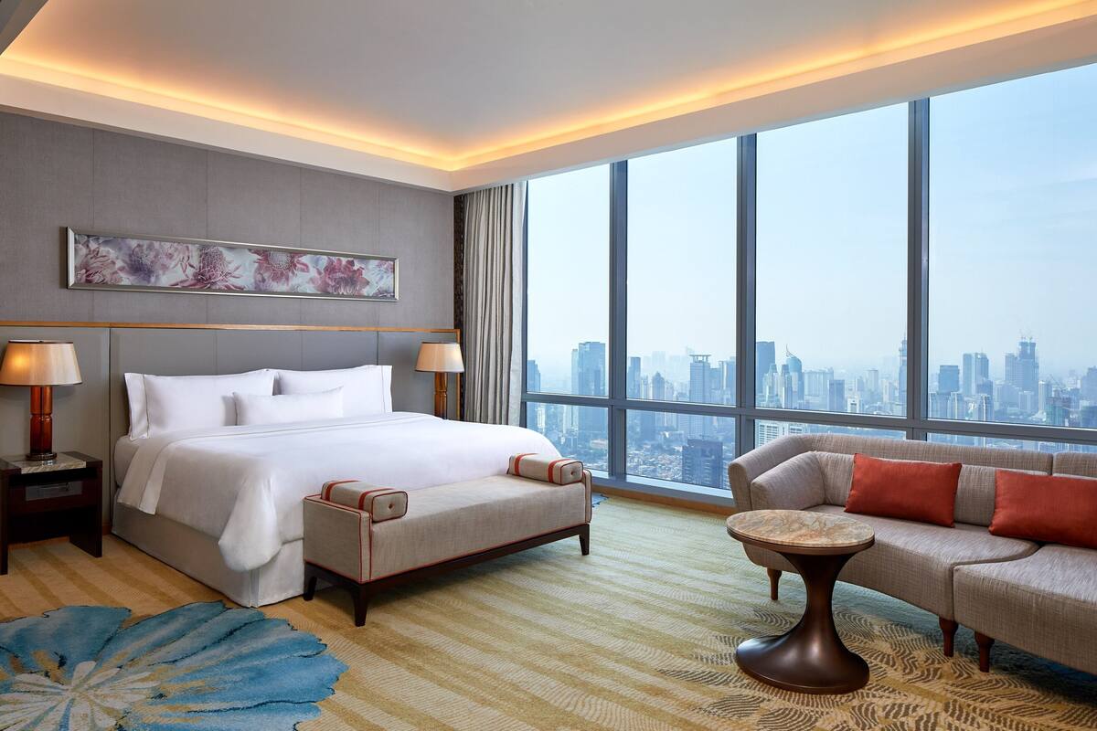 Hotelkamer met een groot bed, een bank, een tafeltje en uitzicht op de stad door grote ramen.