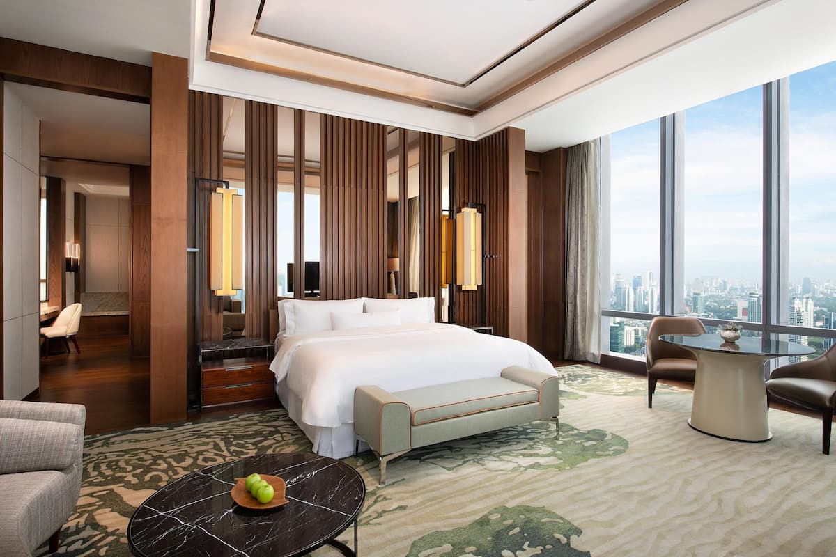 Een moderne hotelkamer met een groot bed, een zithoek met een tafel en stoelen, uitzicht op de stad door grote ramen en een kamerbreed tapijt met dessin.