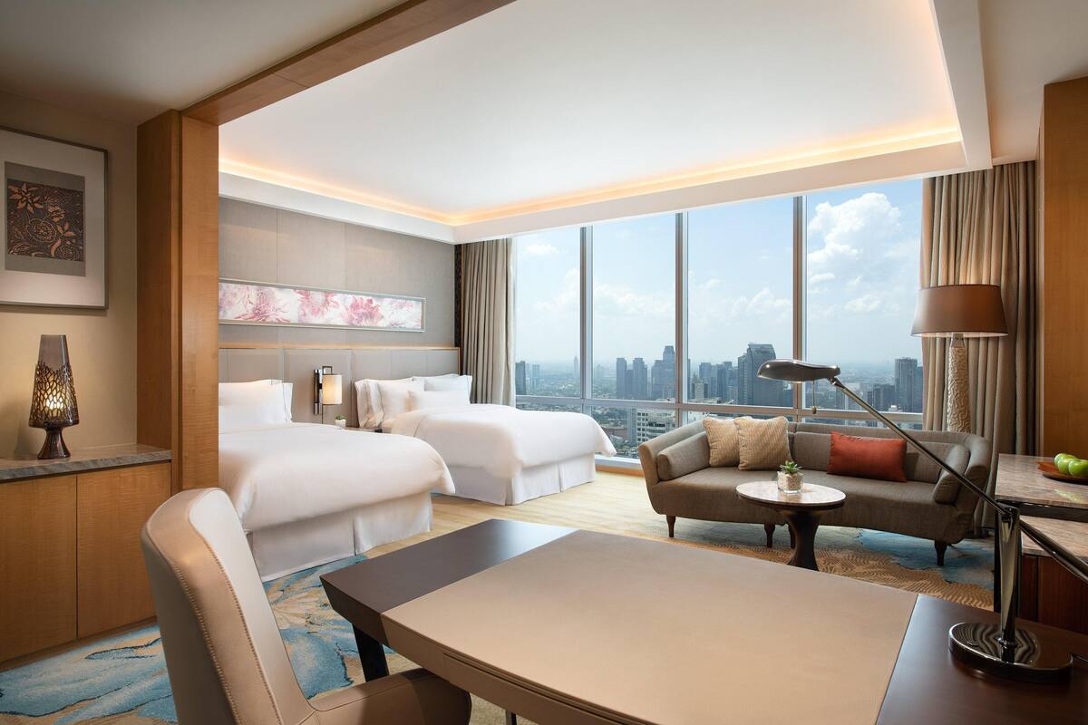 Een hotelkamer met twee bedden, een bank, een eettafel en uitzicht op de stad.
