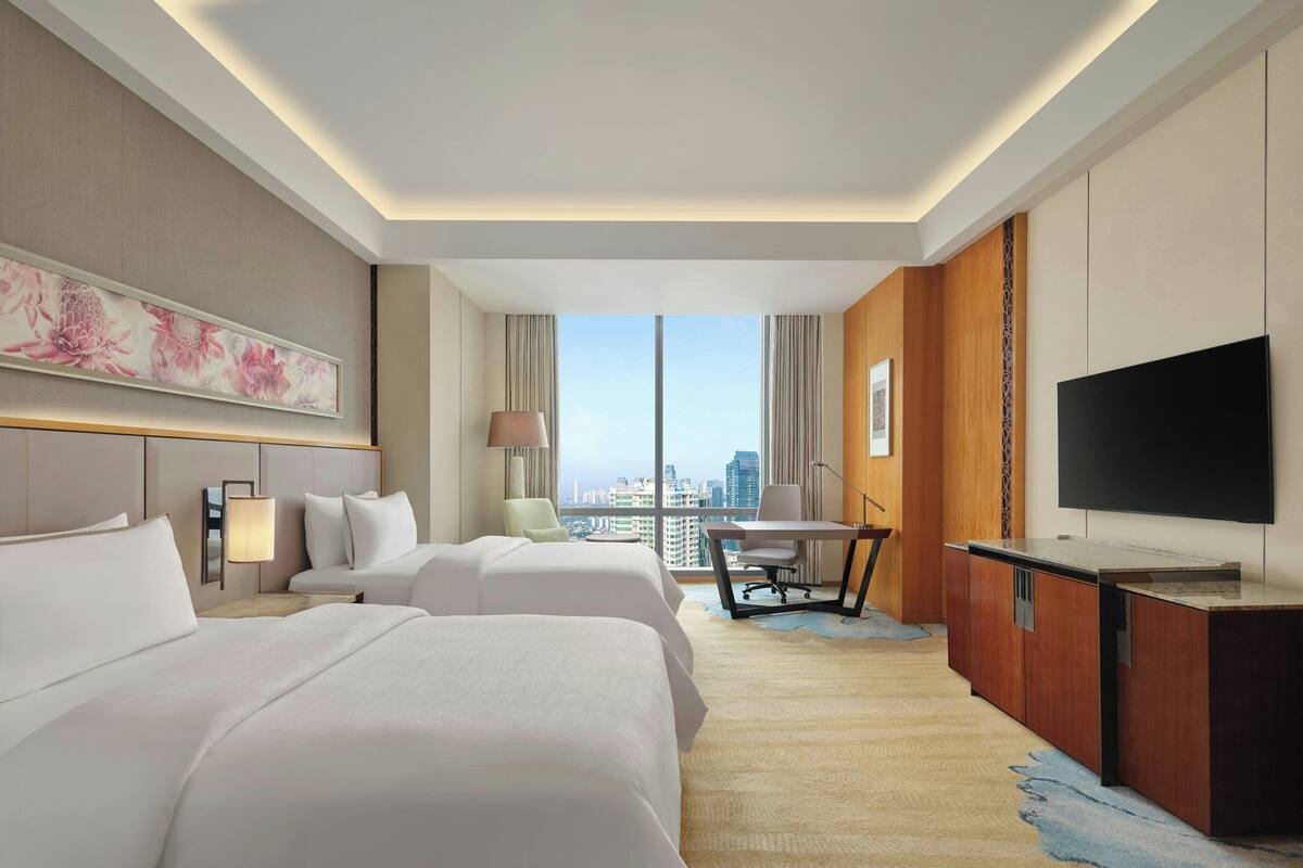 Een hotelkamer met twee bedden, een bureau, een televisie en een groot raam met uitzicht over de stad.
