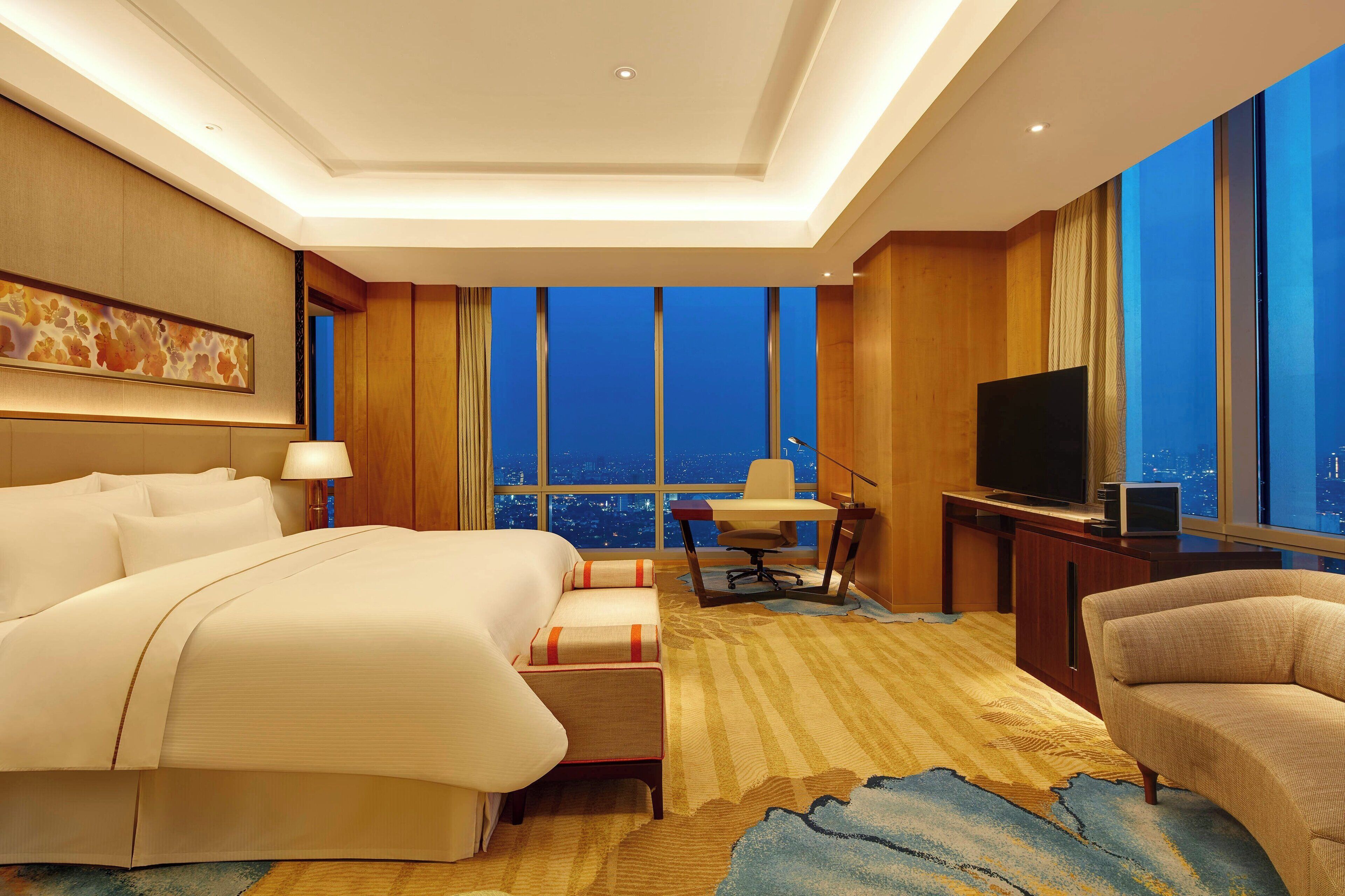 Habitación de hotel con una cama grande, un escritorio con silla, un televisor y vista a la ciudad de noche.