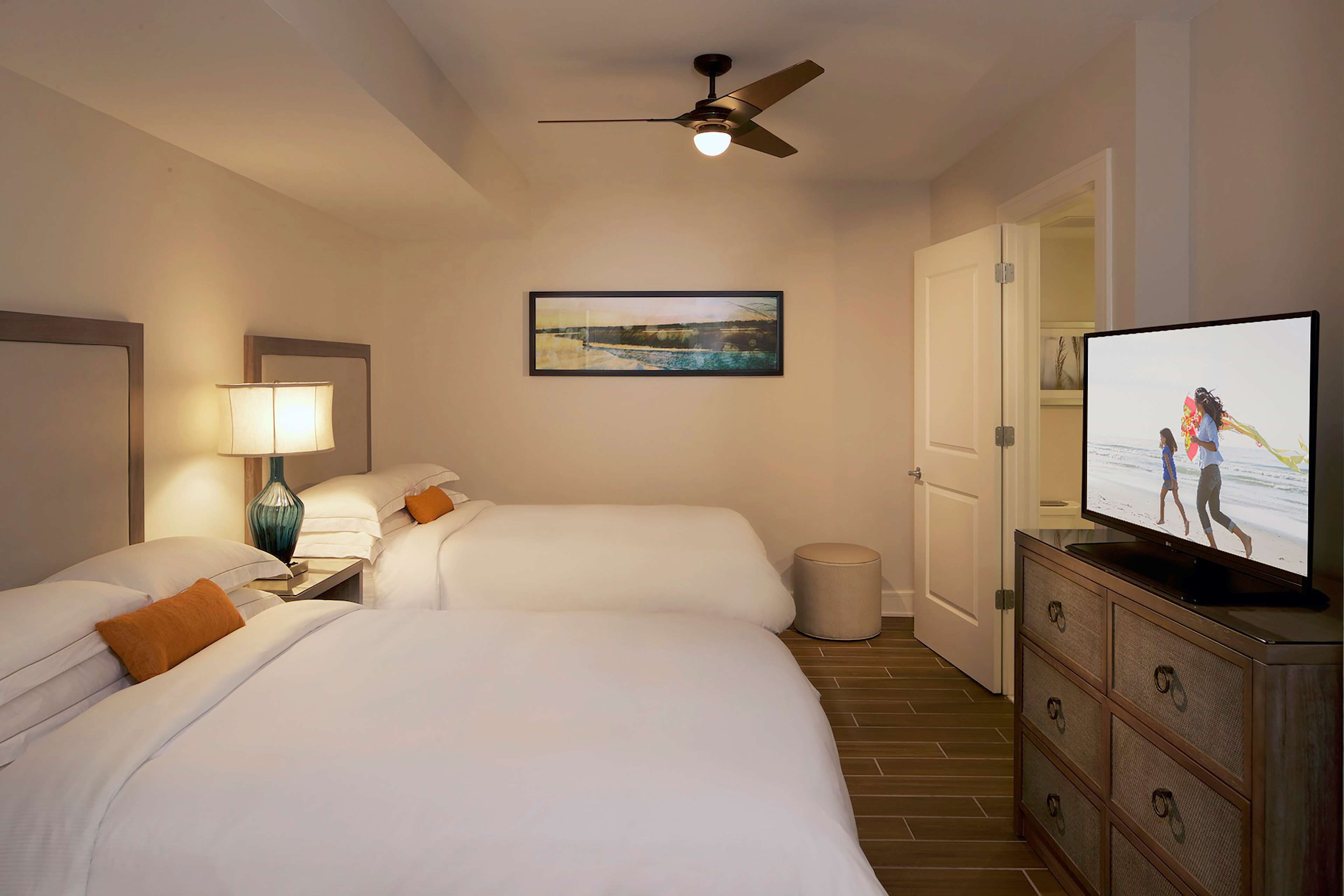 A hotel room with two beds, a ceiling fan, a TV, a lamp, and a framed picture on the wall.