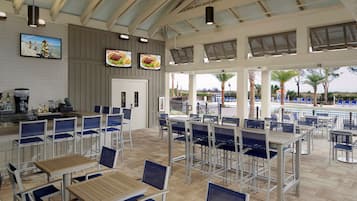 Amplia zona de comedor al aire libre con varias mesas y sillas, una barra y grandes ventanales con vistas a palmeras y una piscina.