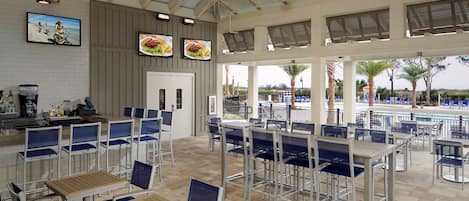 Un vaste espace extérieur aménagé pour les repas, avec de nombreuses tables et chaises, un comptoir de bar et de grandes fenêtres offrant une vue sur les palmiers et une piscine.