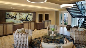 A spacious lobby with a modern reception desk, comfortable seating areas, a staircase, and a large abstract painting on the wall.