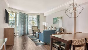 A hotel room with a wooden floor, a dining area with a table and chairs, a sofa, a TV mounted on the wall, and a window with curtains.