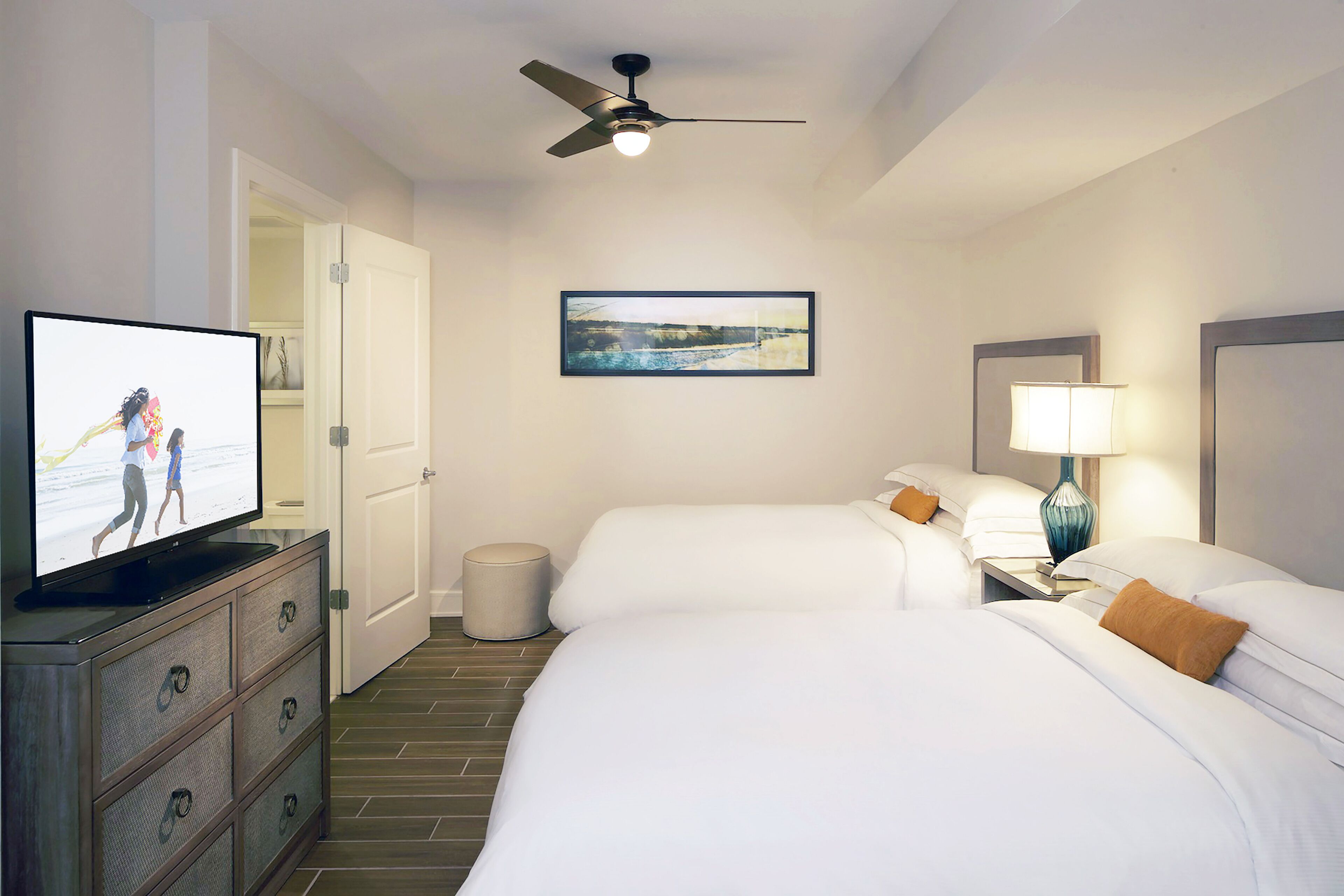 Une chambre d’hôtel avec deux lits, une télévision, un ventilateur de plafond, une poubelle, un tableau encadré au mur et une table de chevet avec une lampe.
