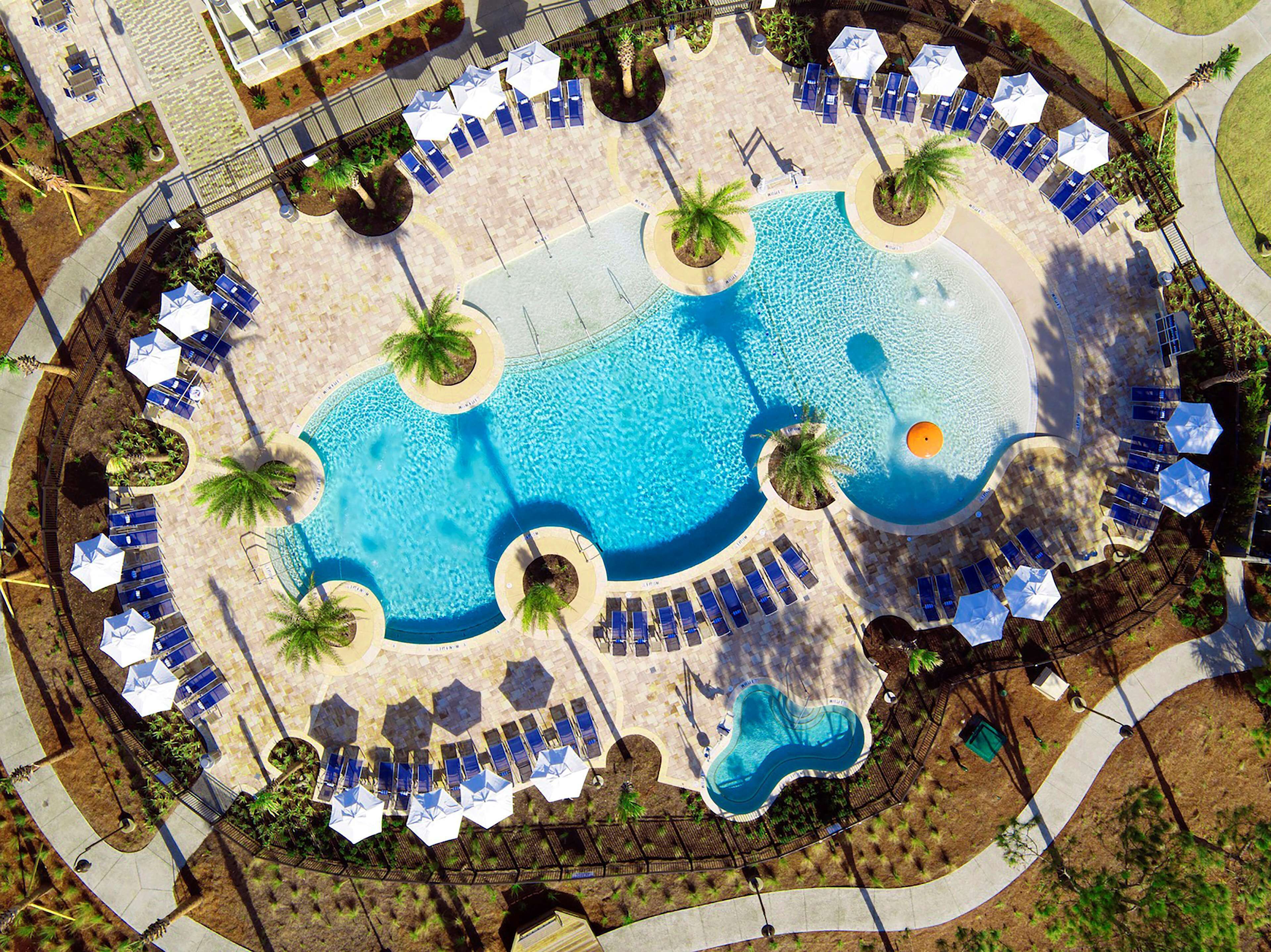 An aerial view of a resort with a central swimming pool, sun loungers, and palm trees.