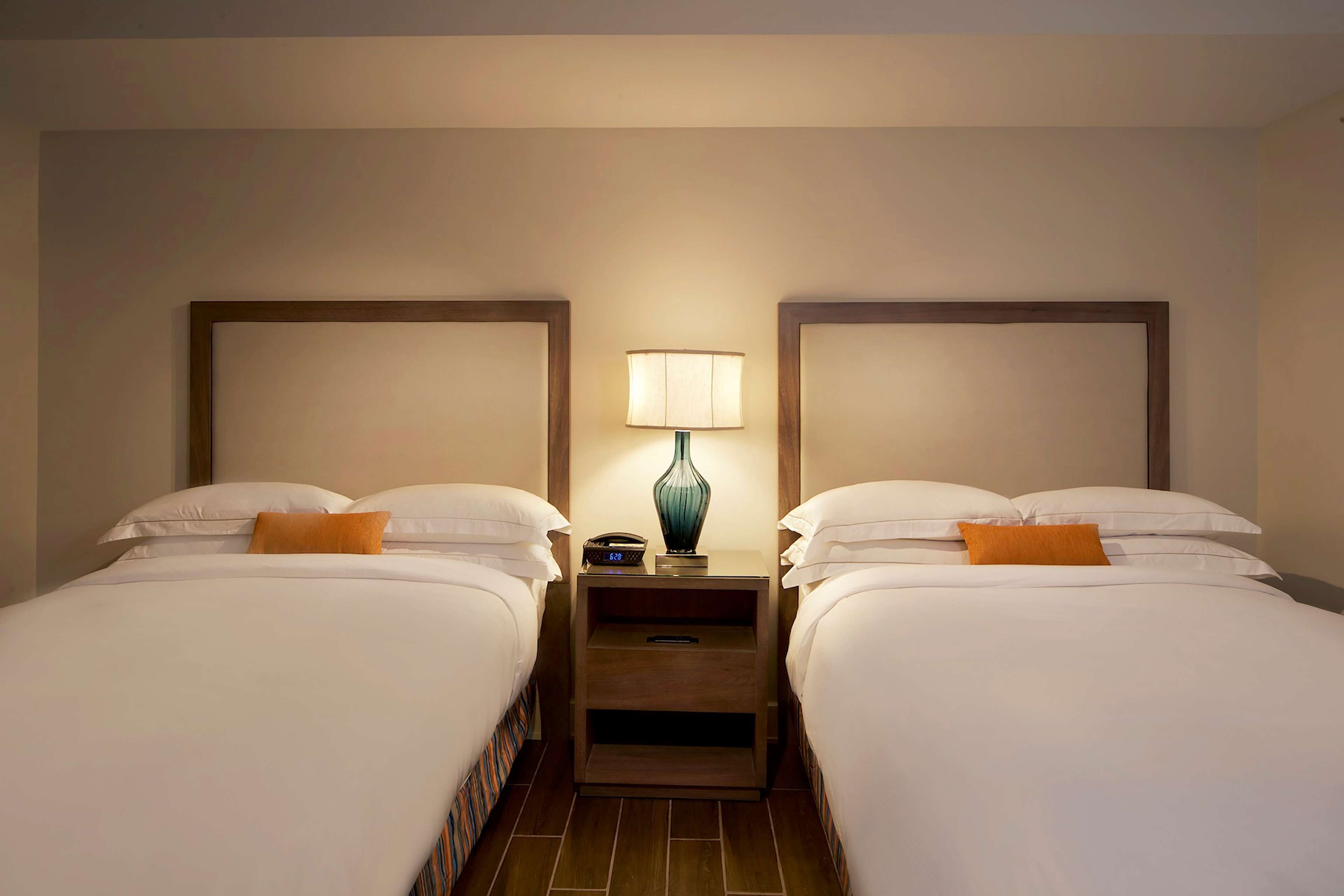 Une chambre d’hôtel avec deux lits, chacun agrémenté de draps blancs et d’un coussin décoratif orange. Une table de chevet avec une lampe se trouve entre les deux lits.