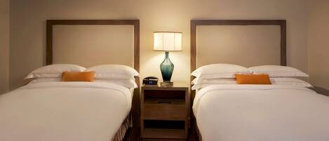 A hotel room with two beds, each with white linens and an orange accent pillow. There is a nightstand with a lamp between the beds.