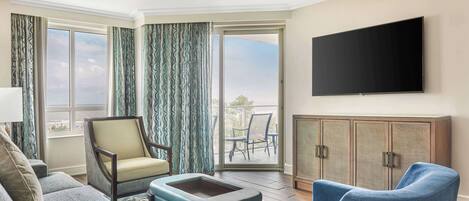 A hotel room with a sofa, armchair, ottoman, a flat-screen TV mounted on the wall, a cabinet, and a sliding glass door leading to an outdoor area with a table and chairs.