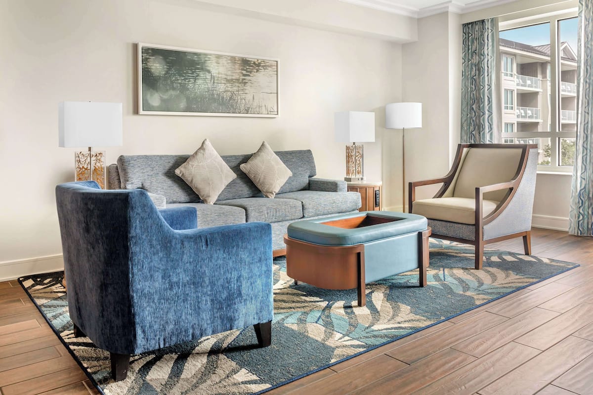 Una sala de estar con un sofá gris, un sillón azul, una mesa de centro de madera y un tapete estampado. Hay dos lámparas de pie y un cuadro en la pared.