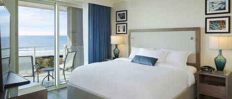 A hotel room with a large bed, a ceiling fan, a view of the ocean, a balcony with chairs, and artwork on the walls.