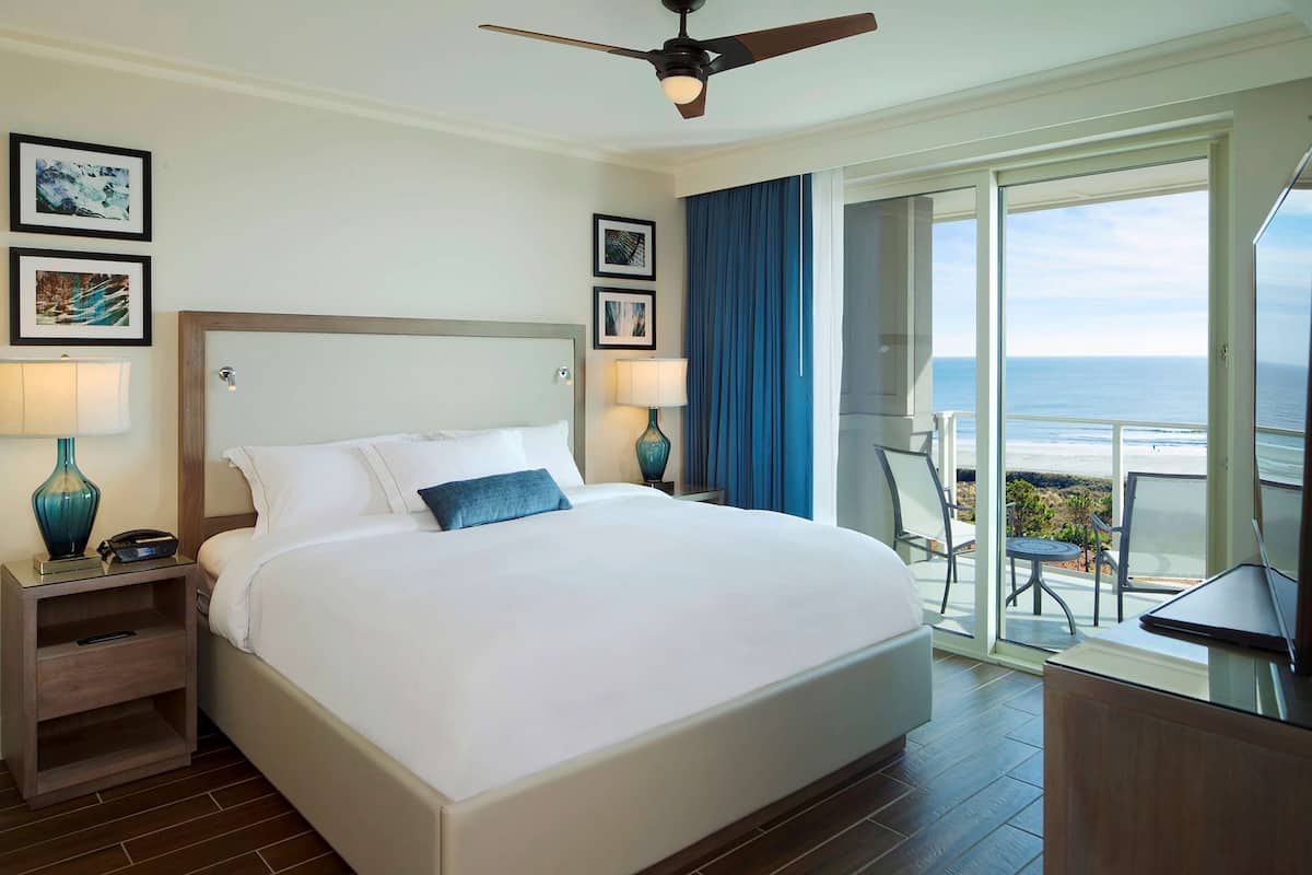 Habitación de hotel con una cama grande, un ventilador de techo, un balcón con vistas al océano y obras de arte en las paredes.