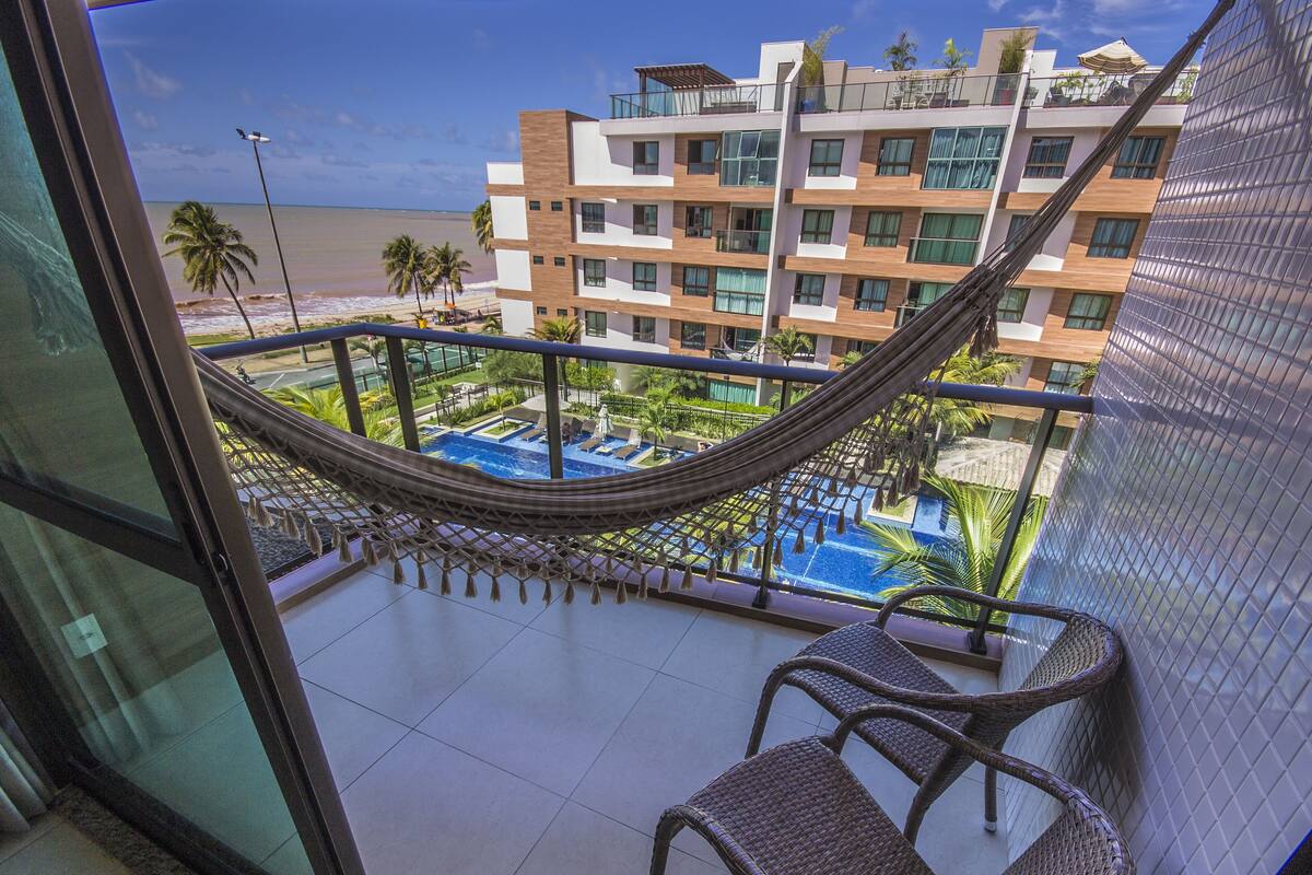 Ein Balkon mit Korbstühlen, einer Hängematte und Blick auf ein Schwimmbecken und einen Strand.
