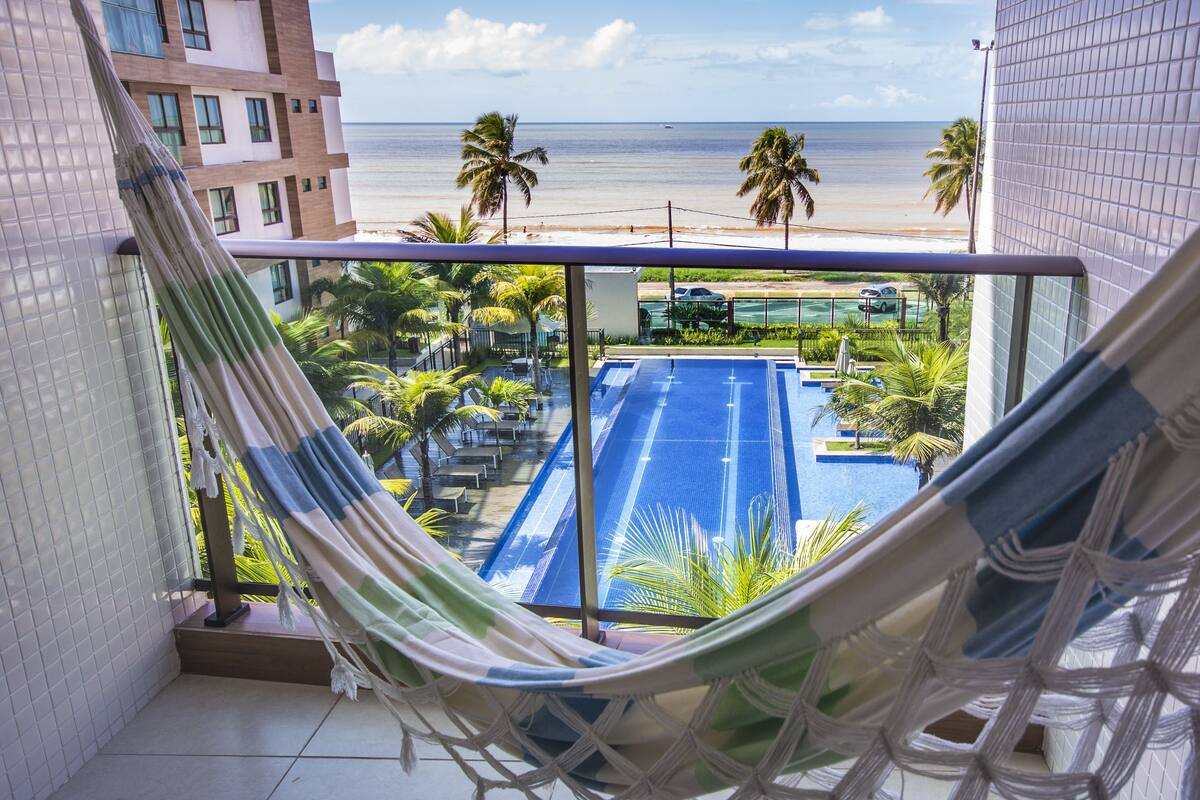 Eine Hängematte auf einem Balkon mit Blick auf einen Swimmingpool, Palmen und einen Strand.