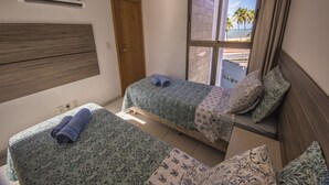 Ein Schlafzimmer mit zwei Betten, einem Holzkopfteil, einem Fenster mit Blick auf Palmen und einen Strand, sowie einer Klimaanlage an der Wand.