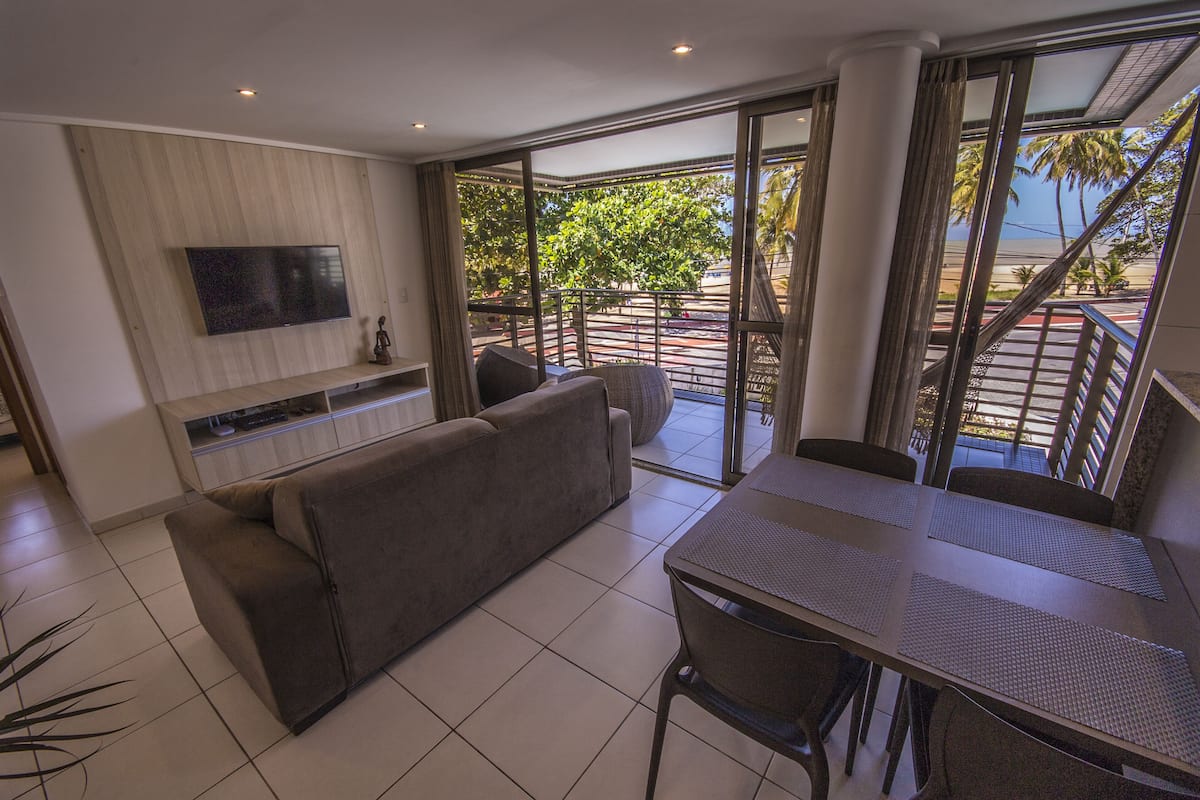 A modern living room with a flat-screen TV, a sofa, a dining table with chairs, and a balcony with a view of palm trees and a beach.