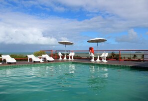 Una piscina con sillas de descanso blancas y sombrillas, una baranda roja y vista al océano.