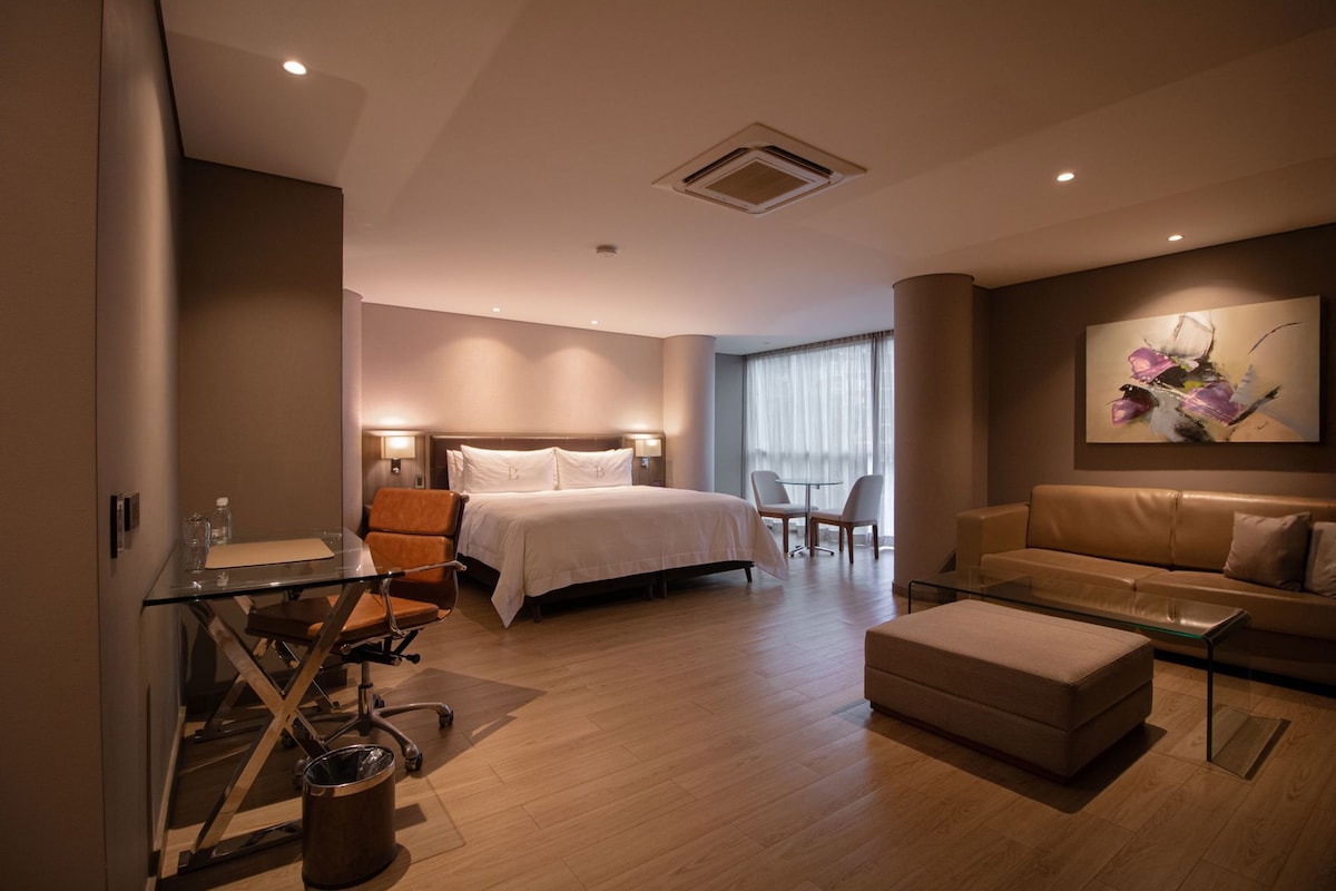 A modern hotel room with a bed, a desk, a chair, a sofa, and a painting on the wall.